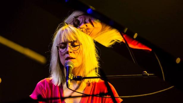 Eine blonde Frau mit Brille singt in ein Mikrofon vor einem Klavier.