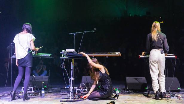 Drei Musikerinnen stehen auf einer Bühne mit Keyboards und technischem Equipment.