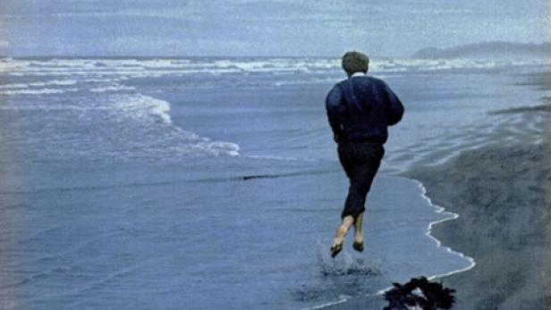 Senator Robert F. Kennedy läuft barfuß mit einem Hund am Strand entlang.