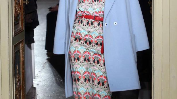 Eine Frau mit einem gemusterten Kleid, einem hellblauen Mantel und roten Schuhen betritt einen Raum.