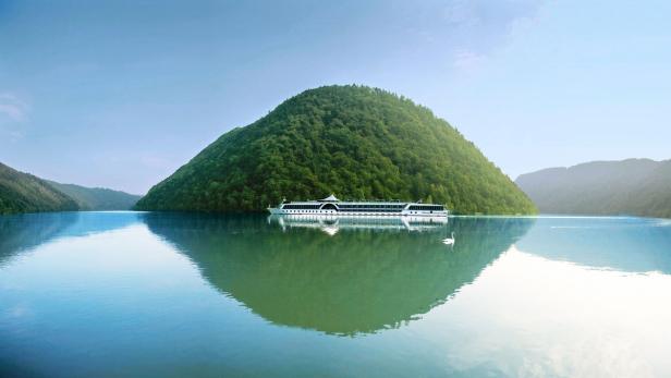 Ein Flusskreuzfahrtschiff fährt auf einem ruhigen See an einem bewaldeten Hügel vorbei.