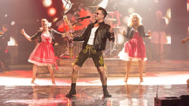 Ein Mann in Lederhose und Lederjacke singt auf einer Bühne mit Tänzerinnen im Dirndl.