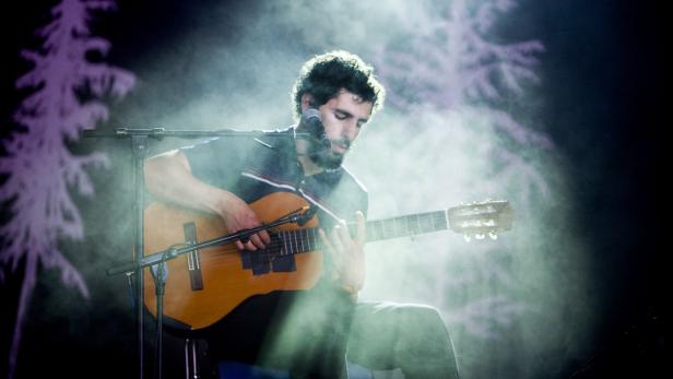 Ein Mann mit Bart spielt Gitarre auf einer Bühne mit Nebel und Lichteffekten.