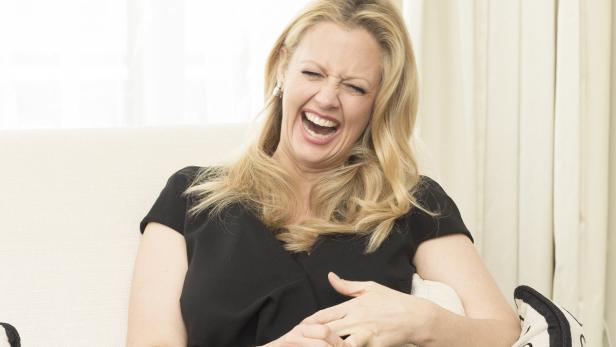 Eine lachende Frau mit blonden Haaren sitzt auf einem Sofa.