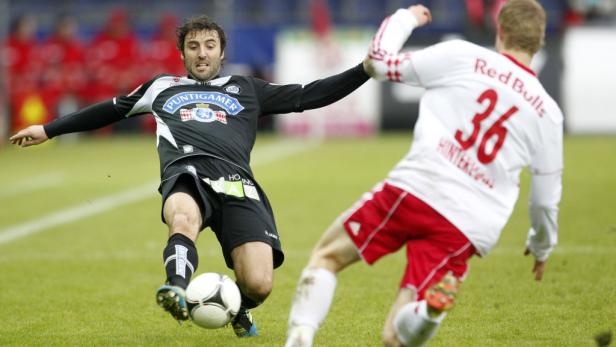 Ein Fußballspieler in Schwarz schießt den Ball, während ein Gegenspieler versucht, ihn zu stoppen.