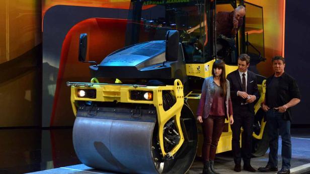 Eine gelbe Straßenwalze mit drei Personen davor auf einer Bühne.