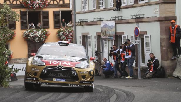Ein goldfarbener Citroën-Rallyewagen fährt durch eine Stadt, umringt von Zuschauern und Fotografen.
