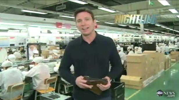 Ein Mann steht in einer Fabrik und hält ein Produkt in den Händen. Im Hintergrund arbeiten Menschen an einer Produktionslinie.