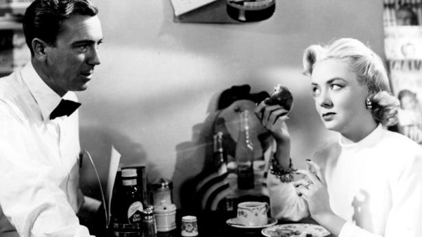 Ein Mann im Smoking und eine blonde Frau sitzen an einem Tisch in einem Diner.