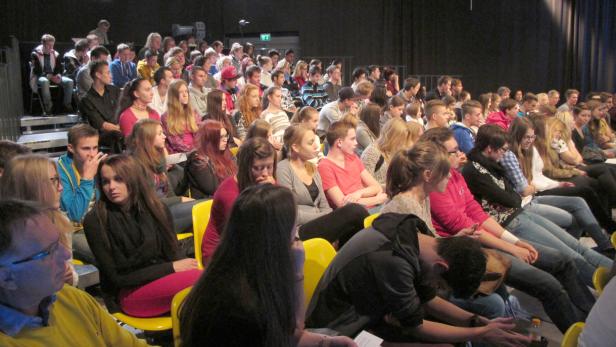 Ein Publikum sitzt auf gelben Stühlen in einem dunklen Raum.