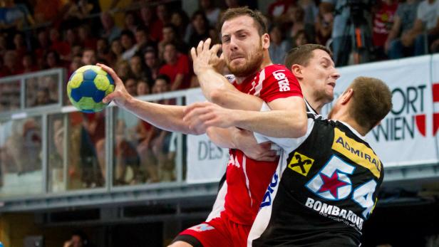 Ein Handballspieler im roten Trikot wird von einem Gegenspieler attackiert, während er zum Wurf ansetzt.