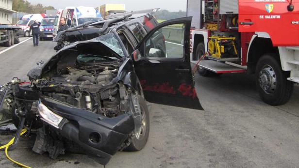 Ein schwer beschädigtes Auto nach einem Unfall, mit Feuerwehr und Rettungsdienst vor Ort.