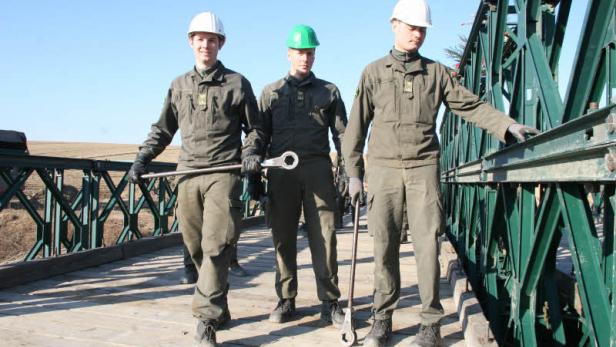 Drei Bauarbeiter in Arbeitskleidung und Helmen stehen auf einer Brücke.