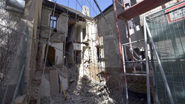 Ein beschädigtes Gebäude, teilweise durch einen Bauzaun verdeckt, mit Trümmern davor.
