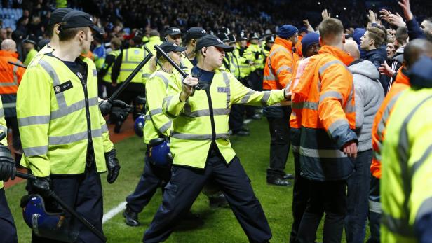 Polizisten in gelben Jacken stehen vor einer Menschenmenge auf einem Fußballfeld.