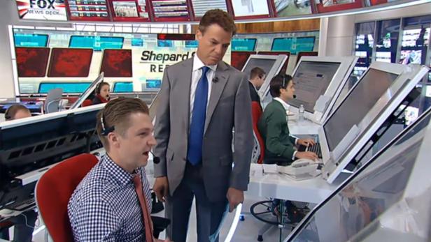 Ein Mann im Anzug steht in einem Nachrichtenstudio und spricht mit einem Mitarbeiter.