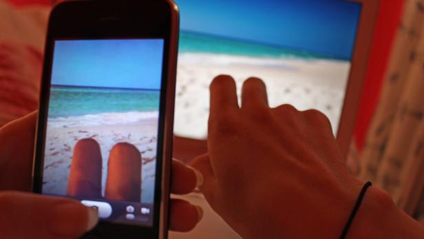 Eine Person fotografiert mit einem Smartphone ein Strandbild auf einem Laptopbildschirm.