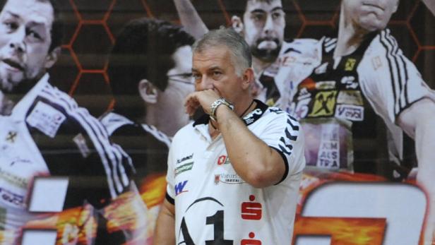 Ein Handballtrainer mit der Hand am Mund vor einem Hintergrund mit Handballspielern.