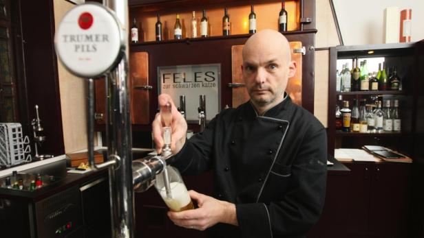 Ein Mann in Kochkleidung zapft ein Trumer Pils in einem Lokal namens „Feles zur schwarzen Katze“.