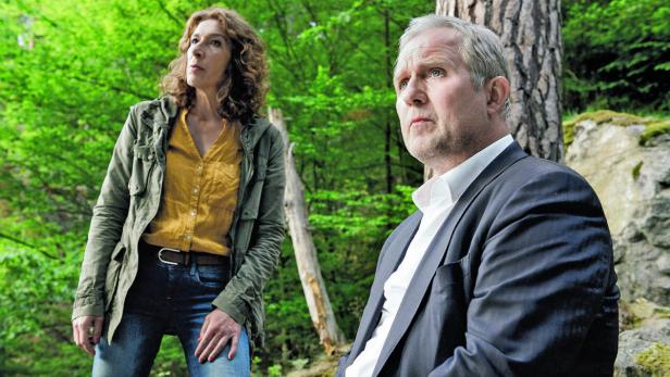 Eine Frau in Jeans und Jacke steht neben einem Mann im Anzug vor einem Wald.