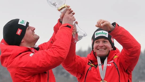 Zwei Männer in roten Jacken und Mützen halten einen Pokal in die Höhe.