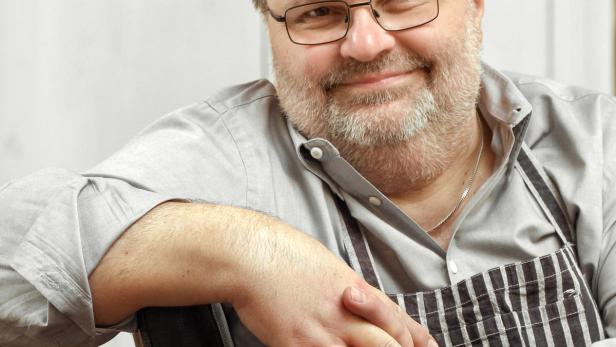 Ein Mann mit Brille und Schürze sitzt auf einem Stuhl und lächelt.