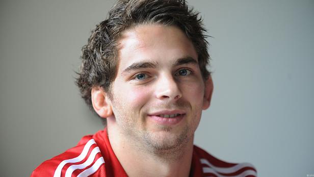 Ein lächelnder junger Mann mit blauen Augen und einem roten Sporttrikot.