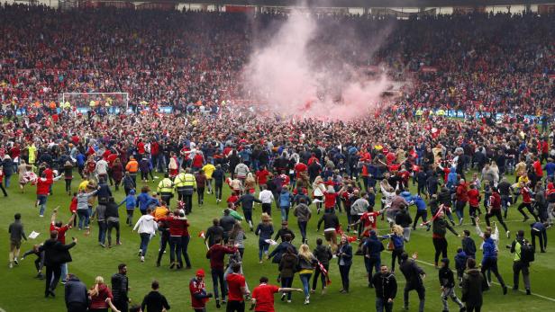 Eine Menschenmenge stürmt nach einem Fußballspiel den Rasen, während rote Rauchbomben gezündet werden.
