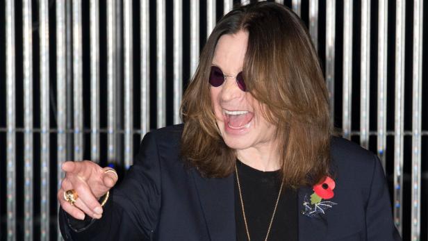 Ozzy Osbourne mit Sonnenbrille gestikuliert vor einem Hintergrund aus vertikalen Lichtstreifen.