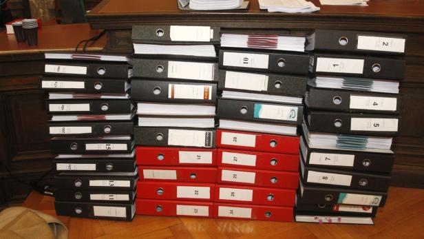 Ein Stapel schwarzer und roter Aktenordner steht auf einem Holzboden.