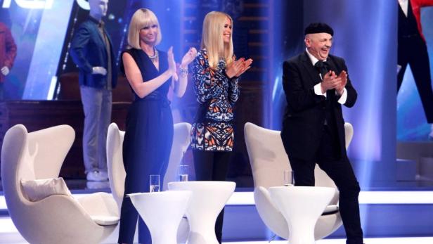 Drei Personen applaudieren auf einer Bühne mit Sesseln und Tischen.