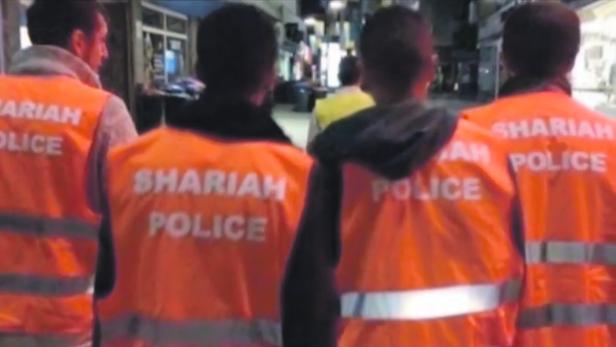 Eine Gruppe von Männern in orangefarbenen Westen mit der Aufschrift „Shariah Police“ auf der Straße.