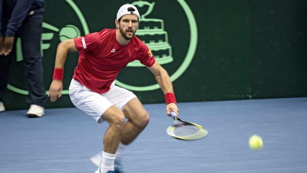 Ein Tennisspieler in rotem Hemd und weißer Hose schlägt einen Ball.