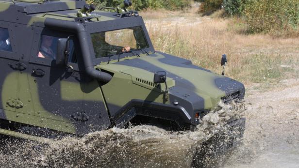 Ein getarntes Militärfahrzeug durchfährt eine Wasserfläche, wobei viel Wasser aufspritzt.