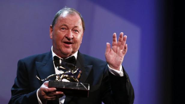 Ein Mann im Anzug hält eine goldene Löwenstatue und winkt.