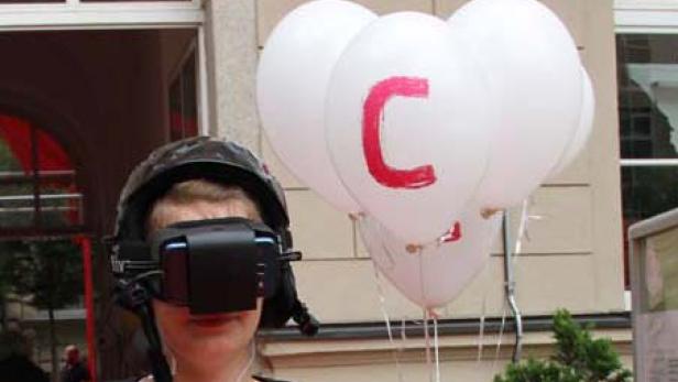 Eine Person mit einem VR-Headset und einem Helm steht neben weißen Ballons mit einem roten „C“.