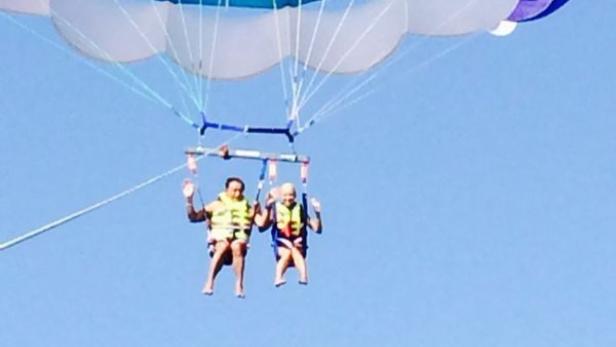 Zwei Personen schweben beim Parasailing mit Schwimmwesten vor blauem Himmel.