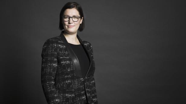 Eine Frau mit Brille und Blazer vor einem grauen Hintergrund.