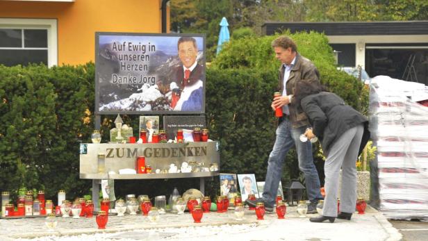 Eine Gedenkstätte für Jörg Haider mit Kerzen und Fotos, besucht von zwei Personen.