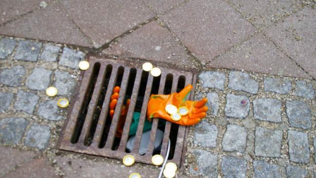 Ein Abwassergitter ist mit einer Arbeitshandschuh und mehreren Euro-Münzen bedeckt.