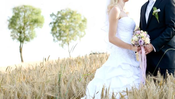 Ein Brautpaar steht in einem Weizenfeld, die Braut hält einen Blumenstrauß.