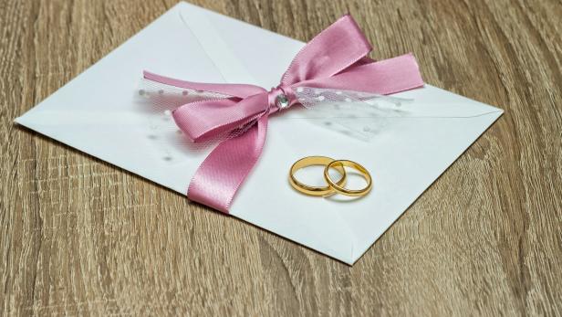 Ein weißer Umschlag mit rosa Schleife und zwei goldenen Eheringen liegt auf einem Holztisch.