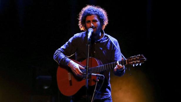 Ein Mann mit lockigem Haar spielt Gitarre und singt in ein Mikrofon auf einer Bühne.