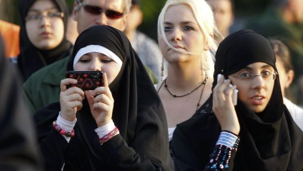 Eine Gruppe von Frauen, einige mit Hijab, nutzen Handys und Kameras in einer Menschenmenge.