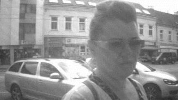 Eine Person mit Sonnenbrille steht vor einer belebten Straße mit geparkten Autos.