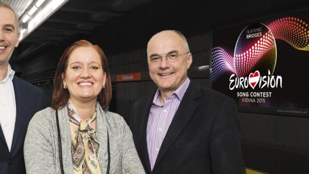Drei Personen stehen vor einem Bildschirm, der für den Eurovision Song Contest 2015 in Wien wirbt.