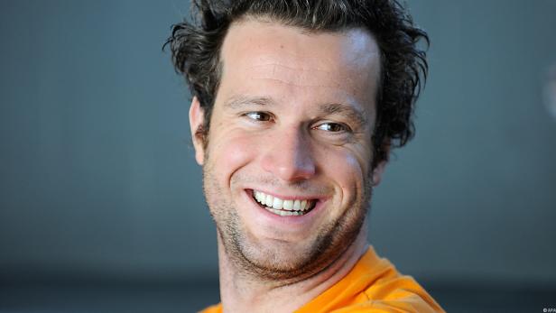 Nahaufnahme eines lächelnden Mannes mit braunen Haaren und einem orangefarbenen T-Shirt.