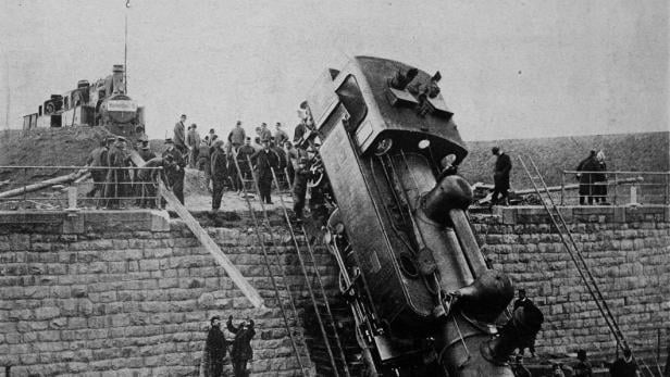 Ein entgleister Zug liegt schräg auf den Gleisen, umgeben von Zuschauern.