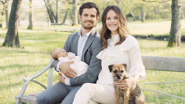 Prinz Carl Philip, Prinzessin Sofia, ihr Baby und ein Hund sitzen auf einer Bank im Freien.