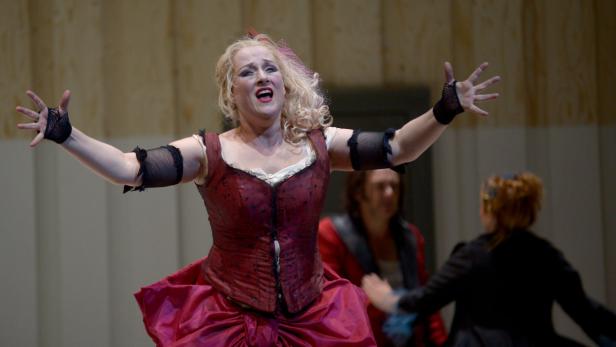 Eine Opernsängerin in einem roten Kleid mit erhobenen Armen auf der Bühne.